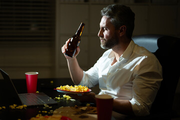 Businessman working late watching online content. Freelancer working late with relaxed mood. Man watching laptop during evening rest. Night office with laptop and snacks. Night office relaxation.