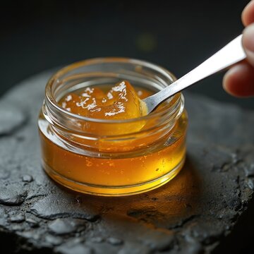 Amber cannabis wax extract sits in glass jar. Metal tool scoops thick golden concentrate. Potent marijuana product for medical use. Legal ganja oil. Closeup detail.