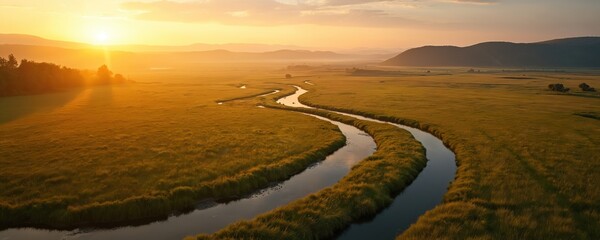 Golden sunset casts long shadows over a wide meadow with a winding river. Rolling hills fade into hazy distance. Lush green grass surrounds calm water reflecting warm sky light.