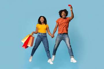 Fototapeta premium Emotional young black couple with shopping bags and credit card holding hands and smiling, jumping up over blue studio background, happy man and woman enjoying seasonal sale, purchasing together