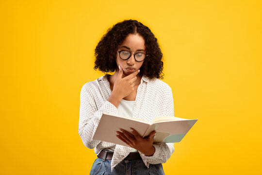 A young woman is standing against a bright yellow wall while reading a book. She has curly hair and wears glasses. She appears to be deep in thought, contemplating the content.