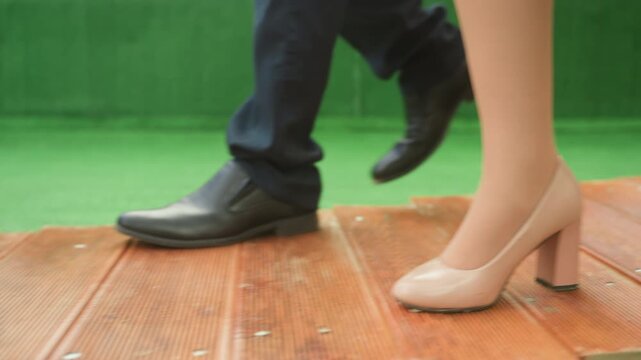 couple walking wooden deck heels shoes, intimate closeup of feet under red table dinner setting, woman wearing nude block heels and man in black leather dress shoes step across weathered planks