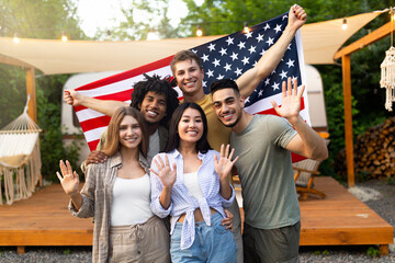 Obraz premium Portrait of multiethnic millennial friends with American flag waving at camera and smiling, posing near RV at camping site. Group of diverse young people celebrating patriotic holiday together outdoor