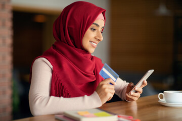 Young muslim lady in hijab using mobile phone and credit card while having coffee break at cafe, buying something online or paying for goods and services via mobile application, copy space