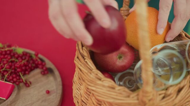 hands arranging apples orange in basket, wicker basket placed on red tablecloth with glass tumblers, bowl of apricots and currants nearby, gentle hands selecting fruit for intimate picnic date, warm