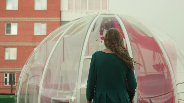 Rooftop igloo date caucasian couple prepare intimate surprise, man adjusts red drapes and hangs floral decoration while woman in green dress follows with wine, transparent dome on terrace, city