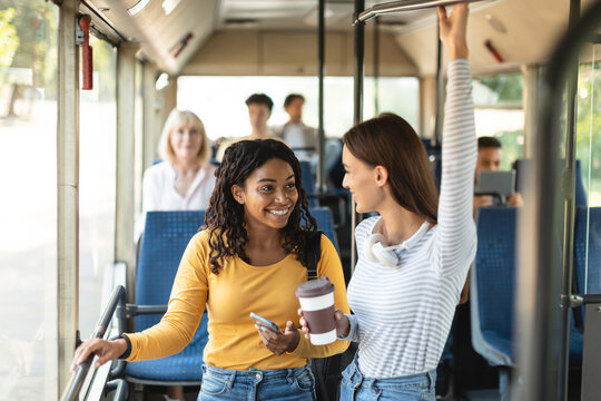 Portrait of two happy young multiethnic ladies standing inside bus and chatting, drinking coffee cup and using cellphone technology, friends commuting together to work or high school university