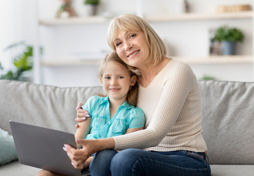 Portrait of smiling senior grandma and cute little kid granddaughter watching cartoons on laptop together, happy older grandmother playing game, sitting on couch hugging and looking at camera - Powered by Adobe