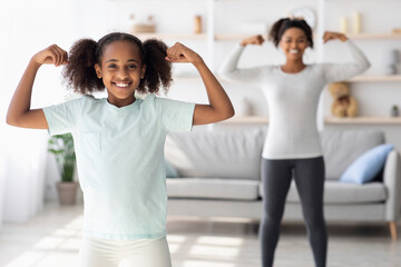 Cheeful teen black girl in sportswear showing her muscles, cute athletic african american kid exercising with mother at home, living room interior. Healthy family lifestyle, sport concept