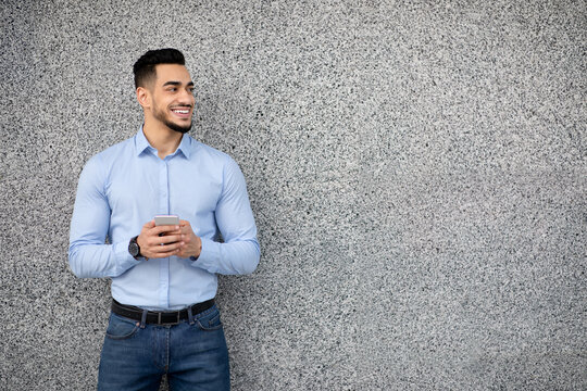 Handsome arab guy in formal wear standing over grey wall, holding cellphone and smiling, chatting with clients or business partners, looking for new business opportunities, copy space, panorama