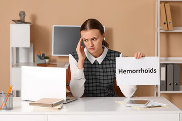 Young businesswoman holding paper with word HEMORRHOIDS at desk in office