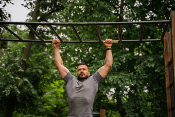 Handsome male athlete in his 40s doing pullups in the park, muscular upper body, hanging on the bar