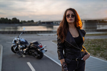 Portrait of very attractive young brunette biker posing in front of her vintage motorcycle, parked by the river 