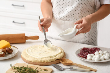 Woman with ingredients spreading tasty white sauce on raw pizza dough in kitchen, closeup