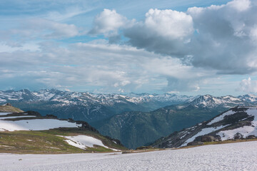 Snowfield on hill top among rocks with view to big snowbound mountain range far away under dramatic cloudy sky. Sunlight and shadows of clouds on forest valley. Sunlit snowy field in high mountains.