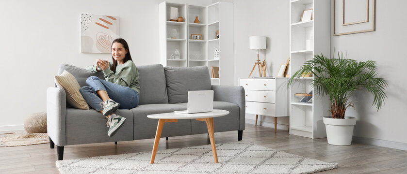 Young woman with cup of coffee sitting on grey sofa in living room