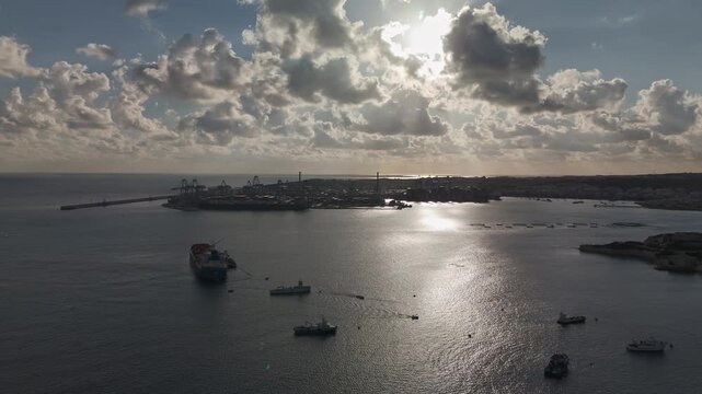 Orbital aerial view around the Malta Freeport in Birżebbuġa, featuring industrial port facilities, ships, and a tranquil Mediterranean seascape beneath a cloudy winter sky.