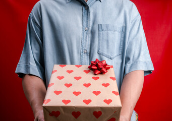 Close-up of man offering a heart-patterned gift box with red ribbon on red background. Ideal for Valentine's Day, birthday, Christmas, or romantic and thoughtful gift-giving concepts.
