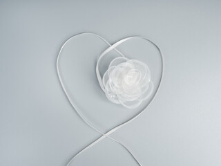 Minimal white ribbon shaped as a heart with delicate fabric flower on soft gray background, symbolizing love, wedding, marriage, romance, purity, commitment, and elegant romantic celebration concept.