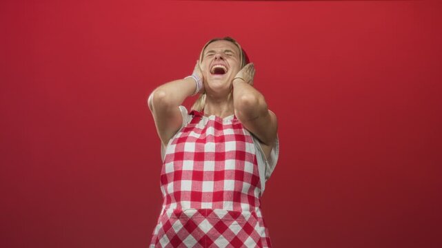 Blonde woman cook in red gingham apron covers ears and winces, hands pressed to ears against red studio backdrop; sensory overload distress.