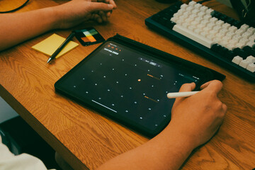 Close up of person hand writing on sticky note while using calendar app on digital tablet at wooden workspace.