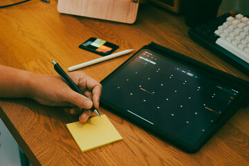 Close up of person hand writing on sticky note while using calendar app on digital tablet at wooden workspace.