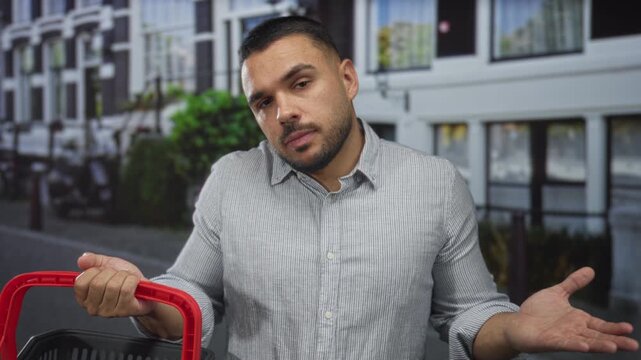Young hispanic man shrugs beside a red recycling container on a busy city street; uncertainty doubt.
