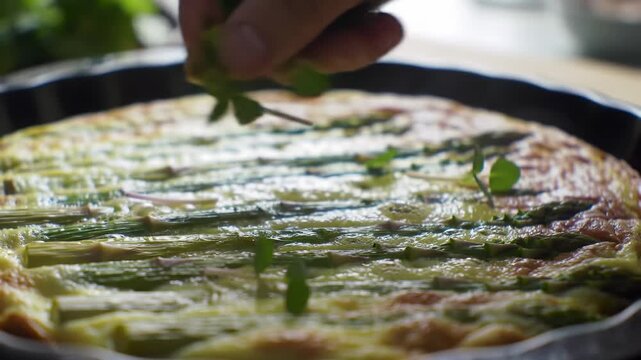 A frittata featuring asparagus. A hand sprinkles topping
