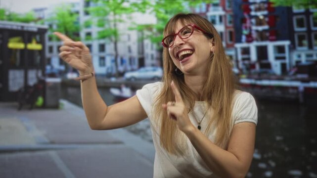 Woman pointing and raising hand showing bare forearm on a street canal in amsterdam, smiling and laughing with red glasses and necklace; joy travel celebration.