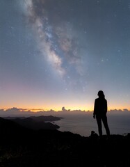 Woman observing the majestic Milky Way over the ocean from a mountain peak at twilight