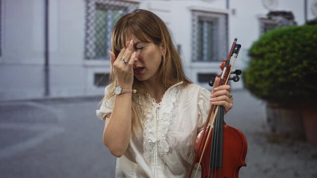 Woman violinist holding violin and bow with hand to forehead in front of building on street; frustration exhaustion.