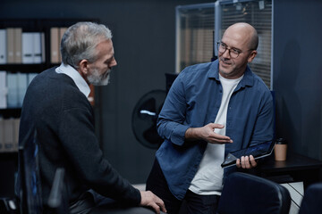 Middle aged Caucasian man sitting and listening while bald man standing and explaining something using digital tablet during business discussion in office
