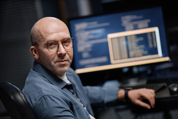Portrait of middle aged Caucasian man with glasses sitting at desk turning toward camera, working on computer with programming code on monitor in modern office setting