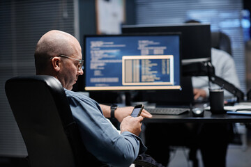 Middle aged Caucasian man sitting at desk using smartphone while working in front of computer monitor displaying code, another visible in background working at separate workstation