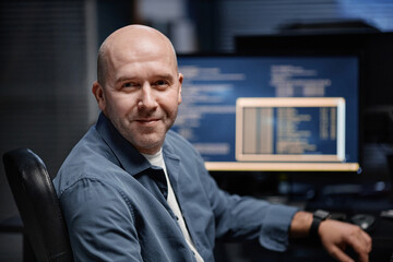 Portrait of middle aged Caucasian man smiling while sitting at desk in front of computer monitor displaying code, showing relaxed expression and looking into camera
