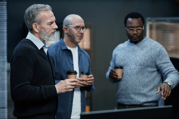 Three middle aged men, including Caucasian and Black individuals, standing together holding disposable coffee cups, engaging in discussion while looking at something off camera in office setting