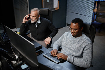Middle aged Caucasian man talking on smartphone while sitting beside young adult Black man working on computer in modern office setting, both focused on tasks, technology visible