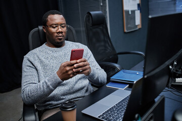 Black middle aged man sitting at desk using smartphone in modern office environment, focusing on device screen with coffee cup, computer monitor and notebooks visible on workspace
