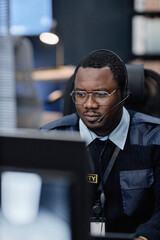 Black middle aged man wearing headset sitting at desk working on computer in office environment, focused expression, security badge visible, performing monitoring or support tasks