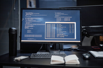 Computer monitor displaying programming code and terminal windows on dark desk, wireless keyboard and mouse placed in front, notebook and reusable coffee cup positioned nearby, modern office setting