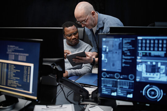 Caucasian man showing digital tablet to young Black man while sitting at workstation with multiple computer monitors, displaying technical data in dark office