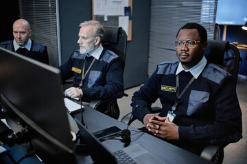 Three middle aged security guards including Black man and two Caucasian men monitoring surveillance screens in control room, sitting at desks with focused expressions, wearing uniforms and badges