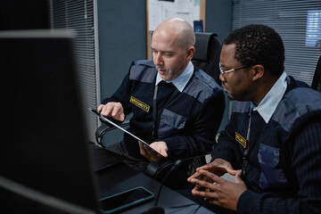 Middle aged Caucasian man and young adult Black man wearing security uniforms sitting at desk, reviewing information on digital tablet in control room with computer monitors visible