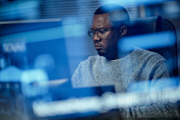 Black man young adult sitting at desk working on computer monitor in modern office, wearing glasses and focusing on screen, technology reflections visible in foreground