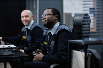 Middle aged Black man and middle aged Caucasian man working together in security control room, Black man holding walkie talkie and sitting at desk, both wearing uniforms and badges