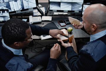 Middle aged Black man and middle aged Caucasian man sitting at security control desk eating sandwiches during break, multiple surveillance monitors and electronic devices visible on desk