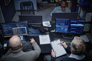 Three middle aged men working in dark office, two Caucasian men analyzing data on multiple computer monitors while Black man observing from background, technology focused workspace