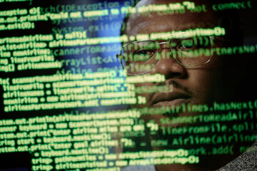 Portrait of Black man wearing glasses looking at computer screen with lines of code reflected on face, focusing on programming or cybersecurity work in technology environment