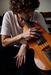 Woman with curly hair plays acoustic bass guitar sitting indoors, wearing a casual white shirt, focusing deeply on creating music with her wooden instrument