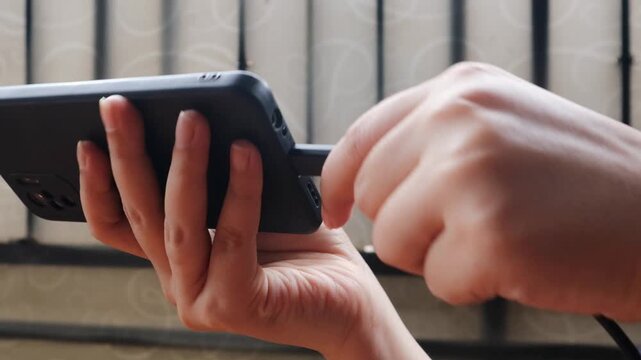 Close-up of an Asian woman's hand plugging a charging cable into a phone. Concept of contemporary technology convenience.